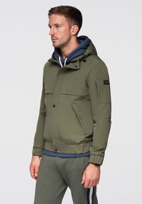 Uomo rivolto verso sinistra che indossa una giacca verde con cappuccio sopra una felpa blu e pantaloni grigi con strisce bianche e blu ai lati su uno sfondo semplice.