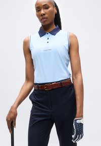 Γυναίκα που φοράει ανοιχτό μπλε χωρίς μανίκια πόλο και σκούρο μπλε παντελόνι, κρατώντας ένα μπαστούνι γκολφ με γάντι γκολφ με σχέδιο στο δεξί της χέρι.