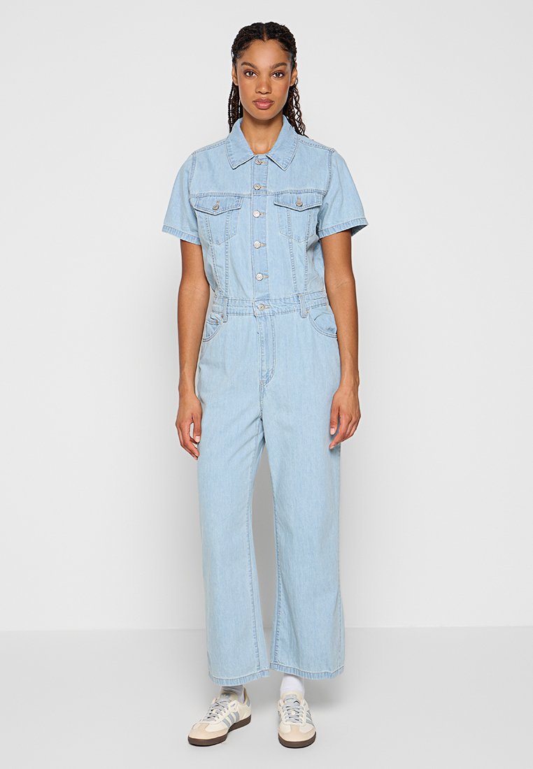 Jumpsuit en denim clair avec des manches courtes, deux poches poitrine, fermeture à boutons et coupe décontractée. Porté avec des baskets de couleur crème.