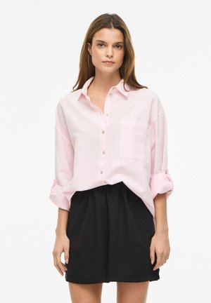 Femme portant une chemise rose clair ample à boutons avec manches retroussées et un short taille haute noir, sur fond blanc.