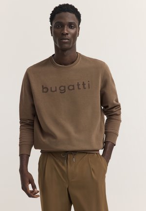 Mann trägt braunes Bugatti-Sweatshirt und passende braune Hose, steht mit einer Hand in der Tasche vor einfarbigem Hintergrund.