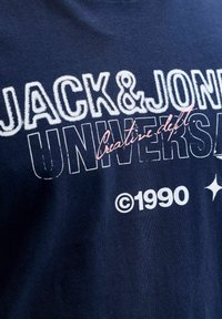 Camiseta de algodón azul marino con "JACK & JONES" en blanco, "UNIVERSITY" en una fuente contorneada y "Creative dept." en rosa, con ©1990 debajo.