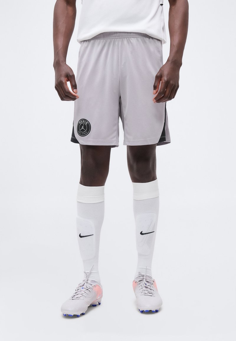 Άνδρας που φοράει γκρι αθλητικές σορτς με το λογότυπο του Παρισιού, λευκές κάλτσες Nike μέχρι το γόνατο και ποδοσφαιρικά παπούτσια, στέκεται μπροστά σε μονόχρωμο φόντο.