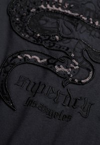 Tejido negro con un bordado detallado de un pulpo estilizado y "Superdry" en hilo negro, con lentejuelas y acentos texturizados.