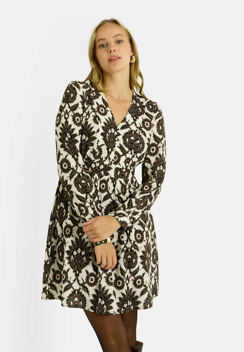 Robe à manches longues blanche avec un motif floral noir, décolleté en v, taille cintrée et jupe évasée. Le mannequin porte un bracelet en or.