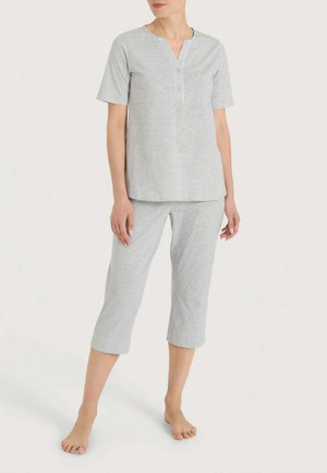 Donna in piedi scalza che indossa una maglia grigio chiaro a maniche corte e pantaloni corti con scollo abbottonato su uno sfondo uniforme.