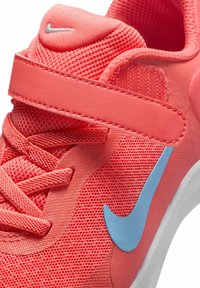 Chaussure de sport Nike en maille corail, avec un swoosh bleu, une tige texturée, un système de fermeture à lacets et sangle, et une semelle rembourrée.
