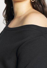 Sudadera negra con escote estilo hombros descubiertos, cuello acanalado y textura suave, con corte holgado y detalles de costuras visibles.