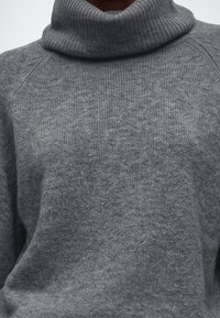 Pull en tricot gris à col roulé avec col côtelé et tissu texturé sur le haut du torse d’une personne.