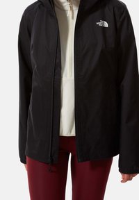 Eine Person trägt eine schwarze The North Face Jacke über einem weißen Reißverschluss-Fleece und dunkellila Leggings und steht mit entspannten Armen.