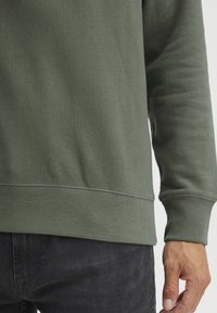 Green sweatshirt with a smooth texture, ribbed cuffs, and a straight hem, paired with dark jeans, showcasing a casual fit.