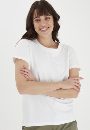 Camiseta de algodón blanca con cuello redondo y mangas cortas, que presenta un corte holgado y una textura suave, llevada por una persona con los brazos cruzados.