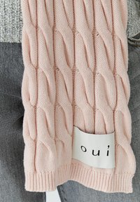 Rosa gestrickter Schal mit einem Kabelmuster, versehen mit einem weißen Etikett, auf dem "oui" in Schwarz gedruckt ist. Die Textur ist weich und hat gerippte Kanten.