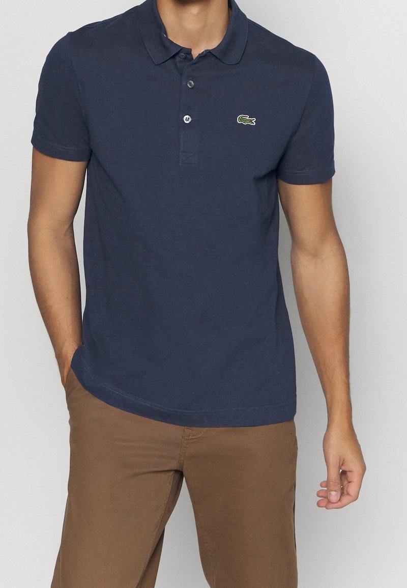 Homme portant un polo bleu marine à manches courtes avec un petit logo vert et un pantalon marron sur un fond uni.