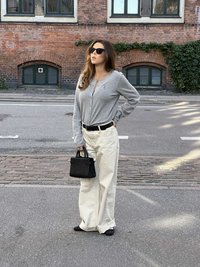 Sivý dlhý sveter na gombíky s dlhými rukávmi, krémové široké nohavice, čierna kabelka a čierne baleríny. Stojí na mestkej ulici s červenými tehlovými budovami za sebou.