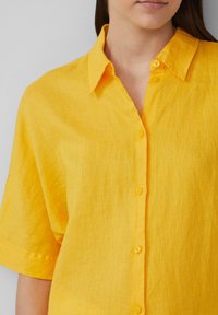 Chemise en lin jaune avec un devant à boutons, des manches courtes et une finition douce et texturée. Équipée d'un col structuré et de boutons assortis.