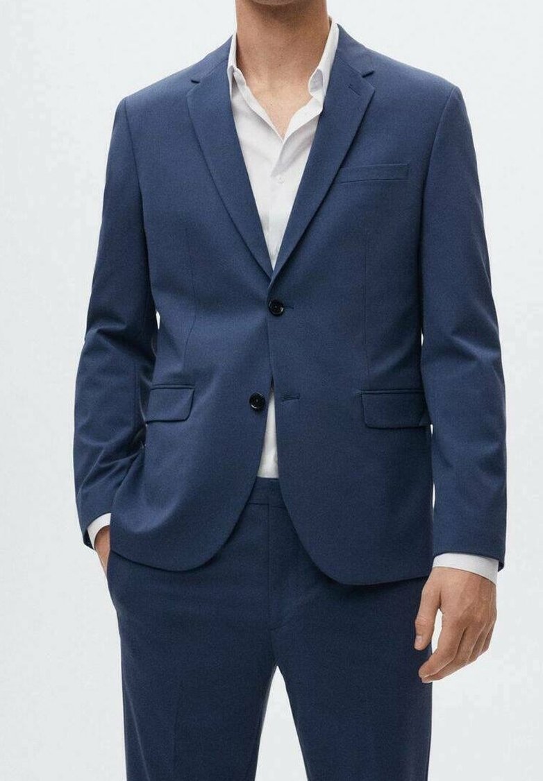 Homme portant un costume bleu marine sur mesure avec des boutons noirs et une chemise blanche, debout avec une main dans la poche.