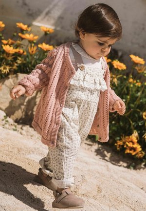 Tout-petit en cardigan rose en maille et salopette fleurie marchant sur un sentier sablonneux avec des fleurs jaunes et des rochers en arrière-plan.