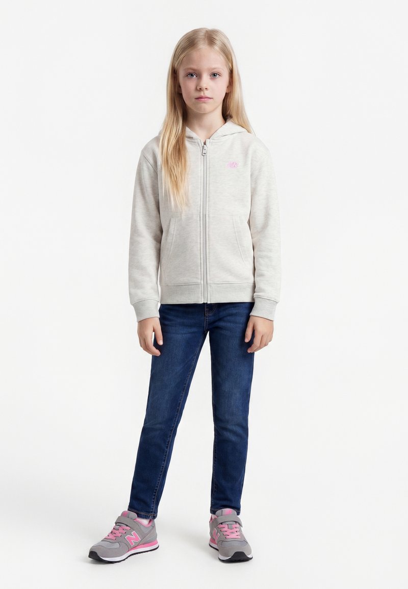 Jeune fille aux longs cheveux blonds portant un sweat à capuche gris clair zippé, un jean bleu foncé et des baskets grises et roses, debout devant un fond blanc.