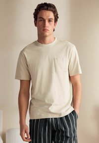 Cremfarbener Kurzarm-T-Shirt mit einer Brusttasche, kombiniert mit gestreiften schwarz-weißen Hosen. Weicher Stoff, lässige Passform.