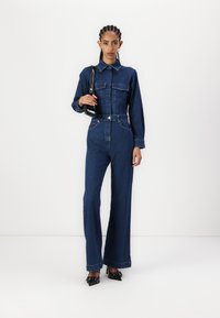 Denim overall in een donkerblauwe tint, met lange mouwen, twee borstzakken, een tailleriem en een brede broekspijp, gecombineerd met zwarte hakken.