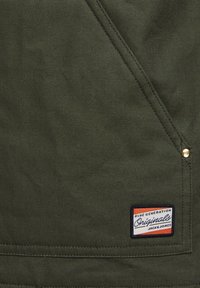 Tissu vert foncé avec détail de couture diagonale, rivet en métal et petite étiquette rectangulaire avec l'inscription "Blue Generation Originals Jack & Jones."