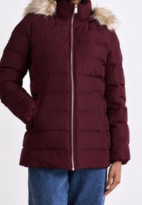 Bordowa kurtka puchowa z wysokim kołnierzem i sztucznym futrem, z zamkiem błyskawicznym z przodu oraz pikowanym wzorem, noszona na niebieskich dżinsach.