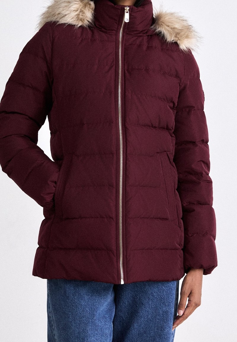 Bordowa kurtka puchowa z wysokim kołnierzem i sztucznym futrem, z zamkiem błyskawicznym z przodu oraz pikowanym wzorem, noszona na niebieskich dżinsach.