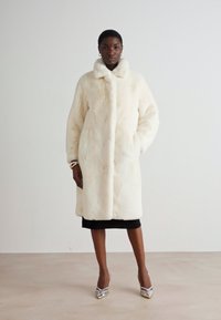 Hvid faux fur frakke med høj krave, lange ærmer og sidelommer. Glat tekstur, midt-lår længde, båret med en sort kjole.