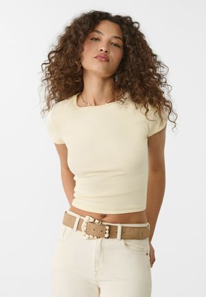Kortärmad, figurnära gul topp i mjukt tyg, kombinerad med högmidjade beige byxor och ett dekorativt bälte med pärldetaljer.