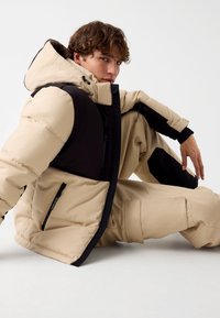 Tans en zwarte geïsoleerde pufferjack met een hoge kraag, ritssluitingen aan de voorzakken en onderscheidende panelen. De stof is soepel met een donzige textuur.