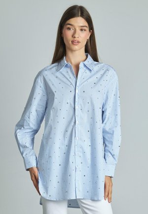 Camicia con bottoni in azzurro chiaro e righe bianche, decorata con piccoli ornamenti argentati. Presenta un colletto e maniche lunghe.
