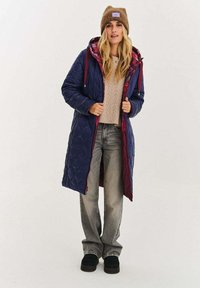 Długi pikowany płaszcz w ciemnym niebieskim kolorze z bordowymi akcentami, wyposażony w kaptur, zapięcie na zamek oraz przednie kieszenie. W komplecie z beżowym swetrem w splot warkocza i szarymi spodniami.