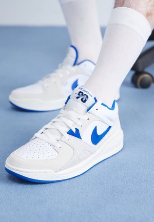 Hvide og blå Air Jordan-sneakers båret med hvide crew-sokker på et blåt tæppe, den ene fod på et hjul under en kontorstol.