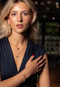 Kultakorut sisältävät kerroksellisia kaulakoruja, joissa on pyöreä riipus ja musta kivi, sekä kultainen rannekoru. Yhdistetty tummansiniseen mekkoon.