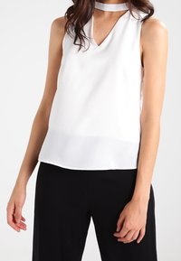 Top blanco sin mangas con escote en V y detalle de collar, confeccionado en una tela suave, con un ajuste holgado y un dobladillo recto.