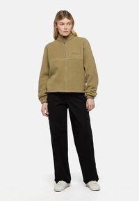 Khaki fleece pullover met een halve rits, met een zachte textuur en subtiele logo; gecombineerd met zwarte wijde broek en witte sneakers.
