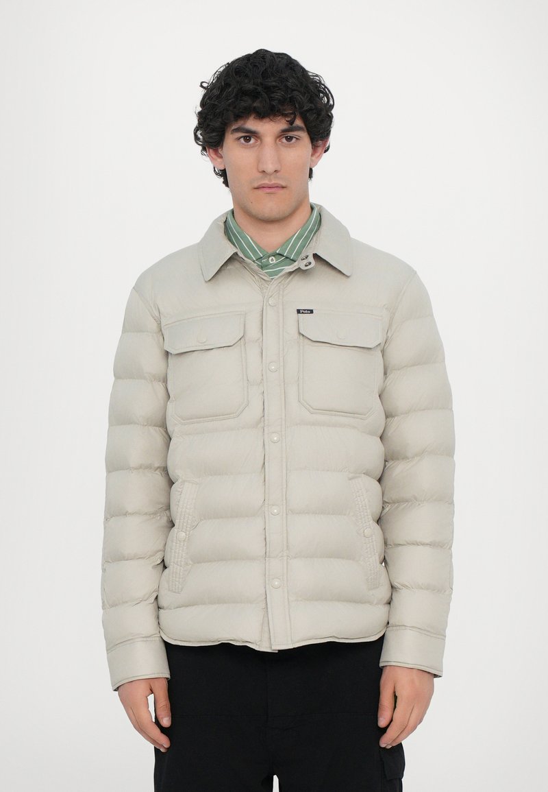 Lys beige puffjakke med knaplukning fortil, to brystlommer og sidelommer. Har en klassisk krave og nylon tekstur.