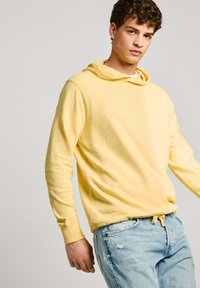 Sudadera con capucha de color amarillo claro, con mangas largas, textura suave y corte relajado. Combinada con unos vaqueros desgastados de color azul claro.