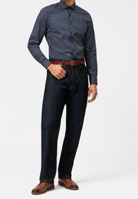 Uomo con camicia a maniche lunghe blu scuro a fantasia, jeans scuri con cintura marrone e scarpe eleganti in pelle marrone, in piedi su uno sfondo bianco.