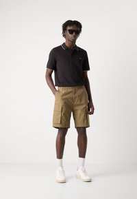 Polo nero con bordi bianchi, pantaloni cargo beige e sneakers bianche. Il modello indossa occhiali da sole scuri e posa su uno sfondo neutro.