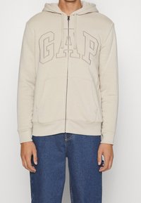 Sweat à capuche beige zippé avec cordon de serrage, logo "GAP" en relief sur la poitrine, poches kangourou, poignets côtelés et intérieur en molleton doux.