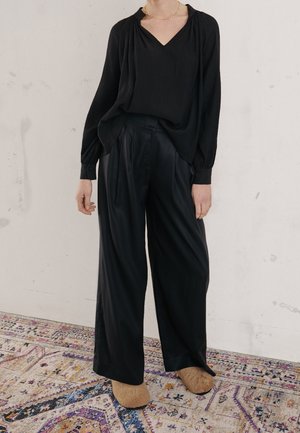 Osoba ubrana w luźną czarną bluzkę i szerokie czarne spodnie stojąca na wzorzystym dywanie, widoczne beżowe kapcie.