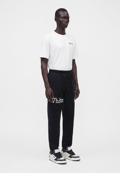Άνδρας που φοράει λευκό t-shirt Off-White, μαύρη φόρμα με λευκό κείμενο "White" και μαύρα-λευκά αθλητικά παπούτσια, στέκεται μπροστά σε απλό φόντο.