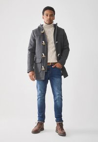 Grijze wollen duffelcoat met knoopsluitingen, grote zakken en een hoge kraag, gedragen over een crèmekleurige kabelgebreide trui en blauwe spijkerbroek. Bruine laarzen.