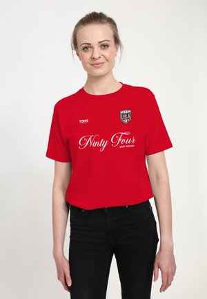 Frau trägt rotes T-Shirt mit USA-Schild-Logo und dem Text „Ninety Four Host Nation“, kombiniert mit schwarzer Hose, stehend vor weißem Hintergrund.