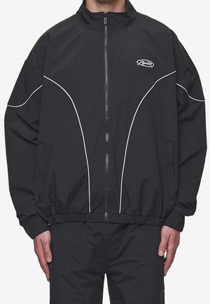 Homme portant une veste noire à fermeture éclair avec des détails en passepoil blanc incurvé et un petit logo sur le haut gauche de la poitrine.