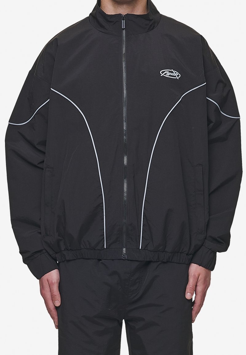 Homme portant une veste noire à fermeture éclair avec des détails en passepoil blanc incurvé et un petit logo sur le haut gauche de la poitrine.