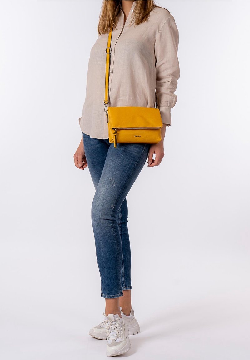 Femme portant une chemise beige, un jean bleu, des baskets blanches, et un sac bandoulière jaune avec un détail de fermeture éclair, debout devant un fond uni.