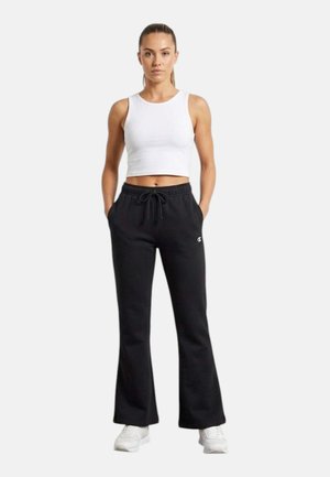 Donna in piedi con le mani in tasca che indossa un top corto bianco senza maniche, pantaloni neri con coulisse e scarpe da ginnastica bianche su sfondo semplice.
